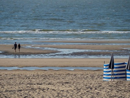 A beach with sand and sea, two people walking along the shore. Blue and white parasols and a quiet