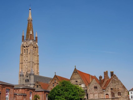 Historic church tower and buildings with tiled roofs, trees and blue sky on a sunny day, Bruges,
