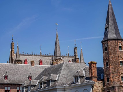 Historic building with many towers and battlements, grey roofs and bricks, under a blue sky,