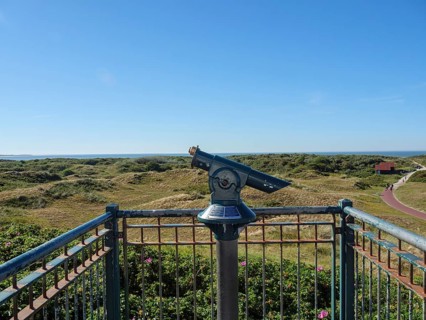 A pair of binoculars on a viewing platform with a view of a vast dune landscape and the sea,