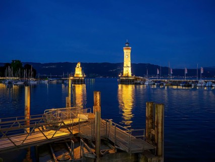 Illuminated harbour at night with lighthouse and lion sculpture reflected in the water, Meersburg,