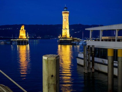 A lighthouse and a statue illuminated at night in the harbour with reflections in the calm water,