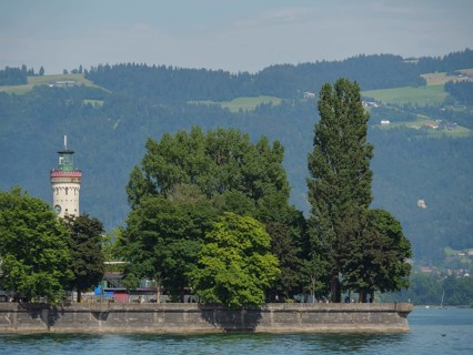 Lighthouse and large trees on the shore with a view of green mountains, Lindau, Lake Constance,