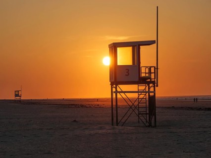 A rescue tower stands on the beach in front of a bright orange sunset, the sea in the background,