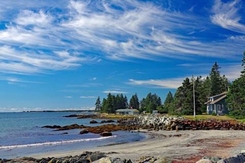 A quiet beach with blue sky, white clouds, sea, sand, rocks, trees and few houses on the coast,