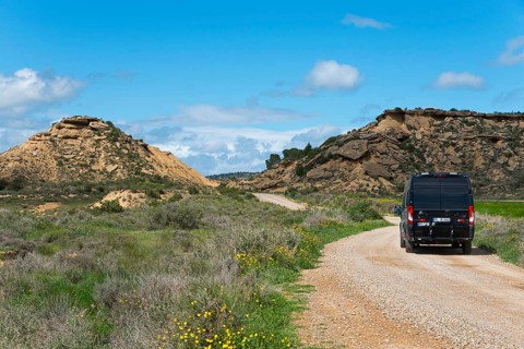 Black van on a gravel road in a hilly, green landscape under a blue sky with clouds, motorhome,