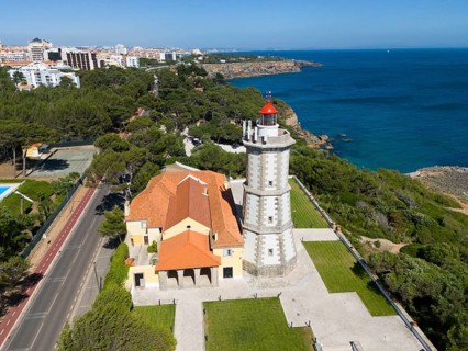 A lighthouse on the coast with a red top and a neighbouring building next to a forest and a paved