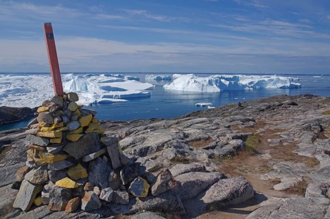 A pile of stones with a flag on rocky terrain in front of an arctic landscape and icebergs in the