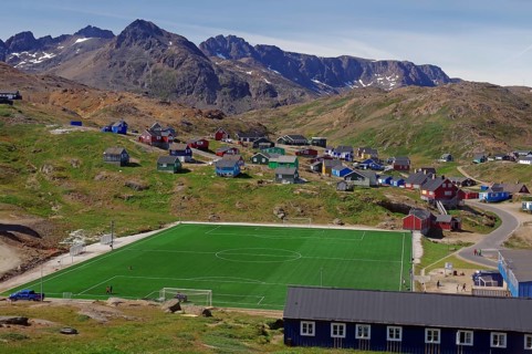 Village with colourful houses and a green football pitch surrounded by mountains and clear skies,