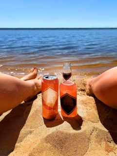 Two people relaxing on a sandy beach with a bottle and can of drink by the water on a sunny day