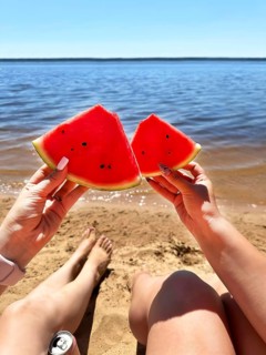 Two hands holding watermelon slices on a sandy beach by a calm lake under a clear sunny sky
