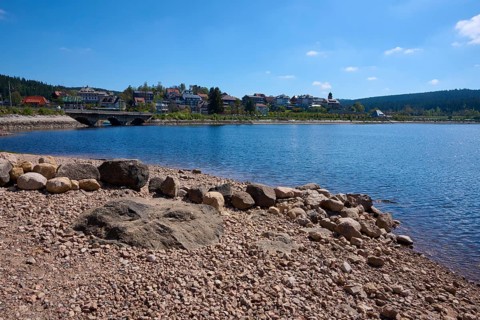 Quiet lake landscape, Schluchsee with pebble beach, bridge and houses on a hill under blue sky,