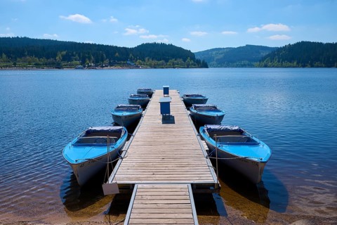 Jetty with moored boats on the Schluchsee, surrounded by mountains and forests, under blue sky,