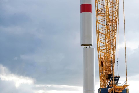 Crane is moving a tower part of a wind turbine onto the first tube guided by people inside,