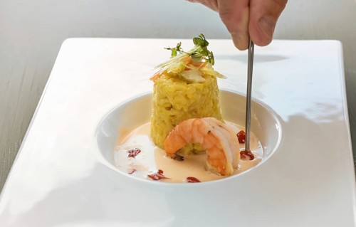 Hand with tweezers garnishing a gourmet dish from saffron risotto with herb topping, a large prawn
