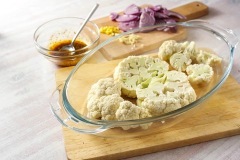 Raw cauliflower slices in a glass casserole, chopped red onion and a spice mix in olive oil,