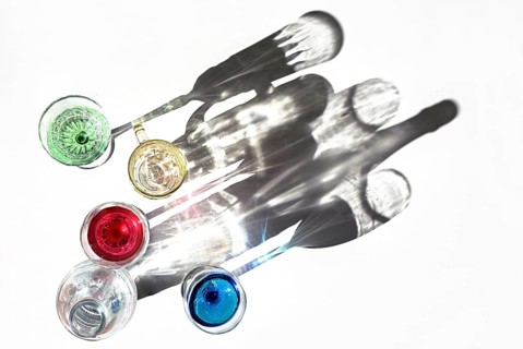 Drinking glasses with colorful liquids and a water bottle are casting shadows on a bright