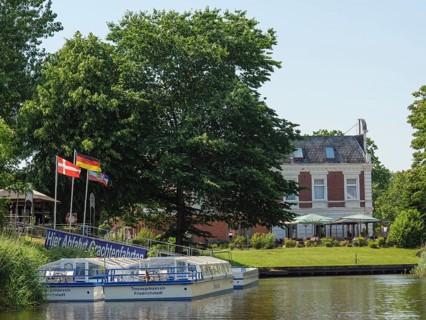 A tourist boat on the canal, surrounded by green trees and a historic villa with flags in the