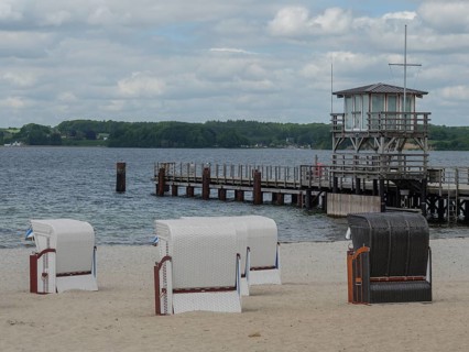 Three beach chairs on a sandy beach, in the background a wooden jetty and the sea under a cloudy