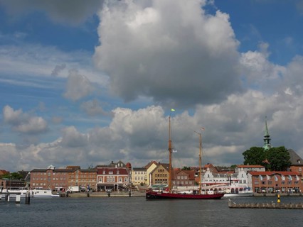 View of a harbour with historic buildings and a sailing ship under a partly cloudy sky, Kappeln,