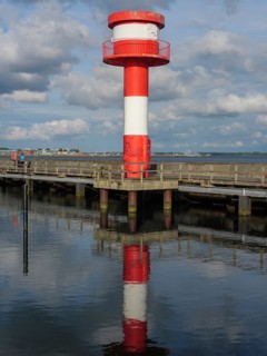 Lighthouse in red and white at the pier, reflecting on calm water under partly cloudy sky,