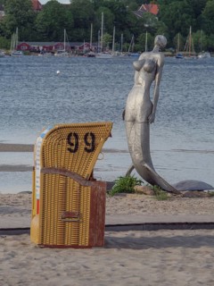 A single beach chair and a statue on a quiet sandy beach overlooking the water under a clear sky,