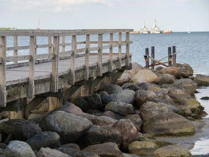 Wooden jetty next to large rocks on the shore with ships in the background, Eckernförde,