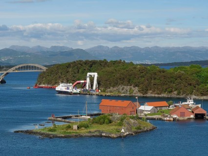 Small island with red buildings and boats, surrounded by the sea. A bridge and mountains are