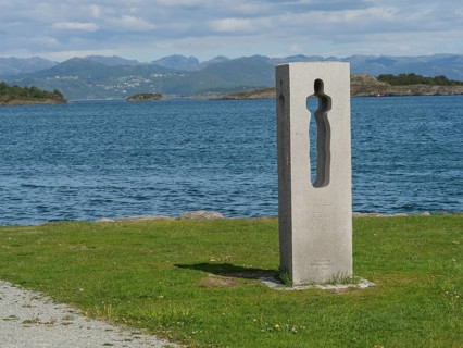 Concrete sculpture with a view of water and mountains, surrounded by green grass and a path in the