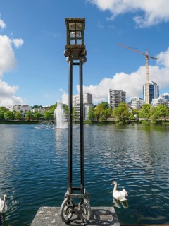 Lamp in the foreground by the lake with a fountain and modern buildings in the background. White