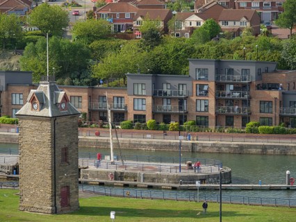 Stone tower on the riverside with modern residential buildings in the background, people walking on