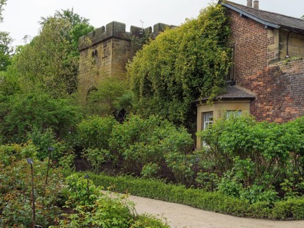 An old fortified tower and neighbouring building surrounded by dense greenery and flowers in a