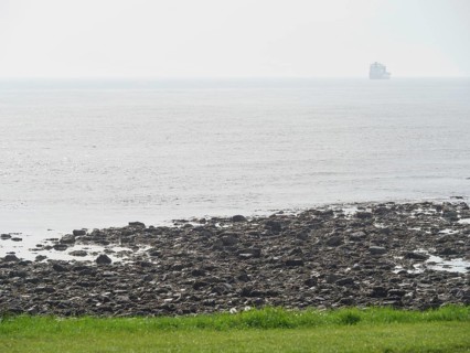 View of a rocky coastline merging into the open sea, a ship can be seen on the horizon in the fog,