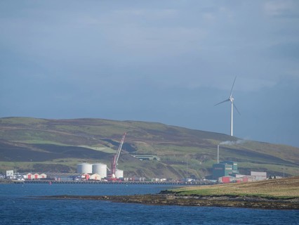 Coastal landscape with green hills, wind turbine and industrial buildings on the horizon, Lerwick,