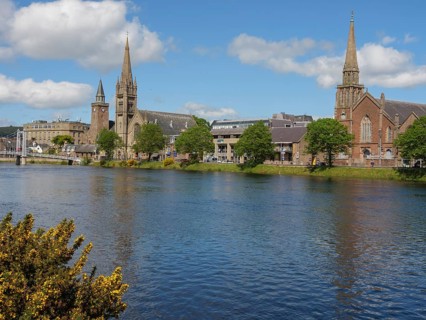 A cosy riverside town with striking church buildings and trees under a partly cloudy sky,