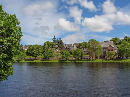 View across a river to a shoreline with trees and historic buildings under a blue sky with some