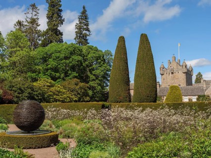 Green garden with hedges, plants and a sculpture, in the background a stone castle under a blue