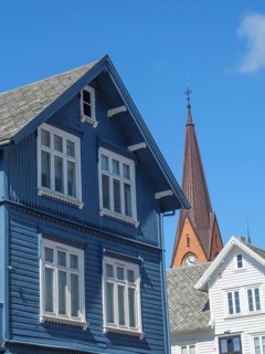 Blue wooden house with white windows in front of a red church tower under a bright blue sky,
