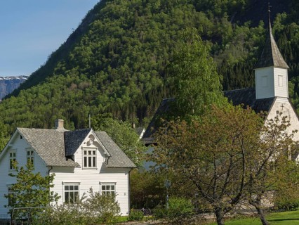 White houses and church in a village surrounded by green trees and mountains under a clear sky,