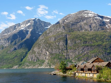 Wooden houses on the lakeshore in front of large, snow-covered mountains under a clear sky,