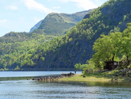 A small cabin on the shore of a clear lake, surrounded by mountains and dense forest, quiet and