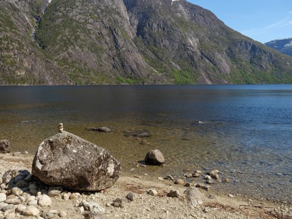 A large boulder lies on a rocky beach with a tranquil fjord and mountains beyond, Eidfjörd, Norway,