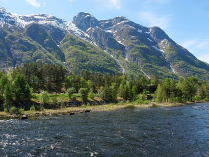 A wide river flows through a wooded landscape with snow-capped mountains in the background,