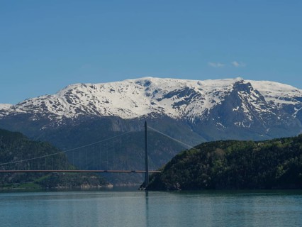 Suspension bridge over a fjord with snow-capped mountains in the background, blue sky and calm