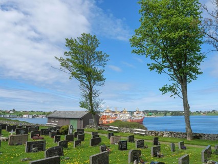 Cemetery with gravestones and trees, behind it a lake and ships under a blue sky, Haugesund, norway