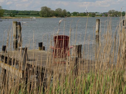 View through reeds to a lake with an old wooden jetty and wind turbines in the distance, Arnis,