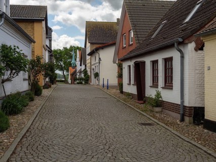 A quiet cobbled street with stylish houses and trees under a cloudy sky, Maasholm,