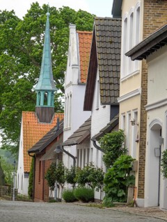 A street with historic buildings and a striking church tower, surrounded by trees and houses with
