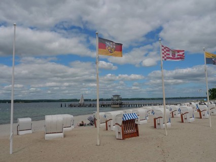 Beach chairs on a sandy beach under a cloudy sky, flags blowing in the wind, sea and jetty in the