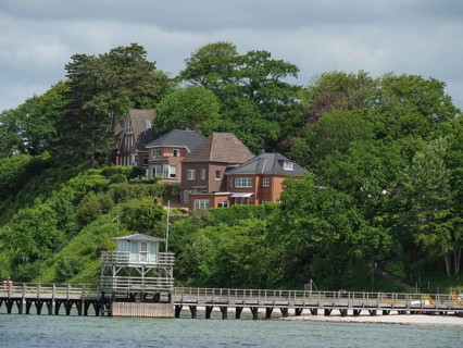 Houses on a green hill, surrounded by trees, with a footbridge leading into the water, under a